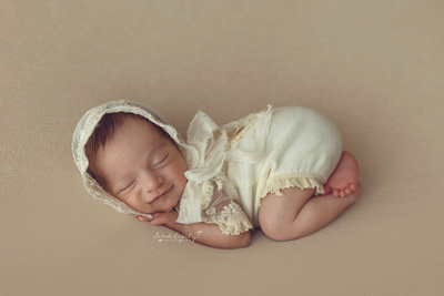 Newborn baby girl on tan background blanket dressed in ivory outfit with matching lace bonnet posed on belly in a NJ photo studio.